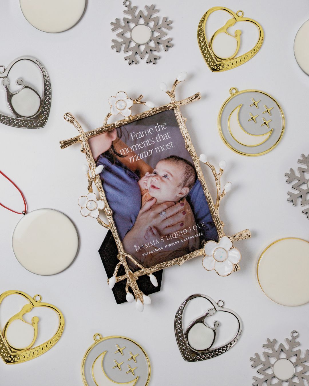 Flatlay of Mamma’s Liquid Love frame and ornaments with the tagline “Frame the moments that matter most,” featuring heart, moon, snowflake, and round breastmilk ornaments.